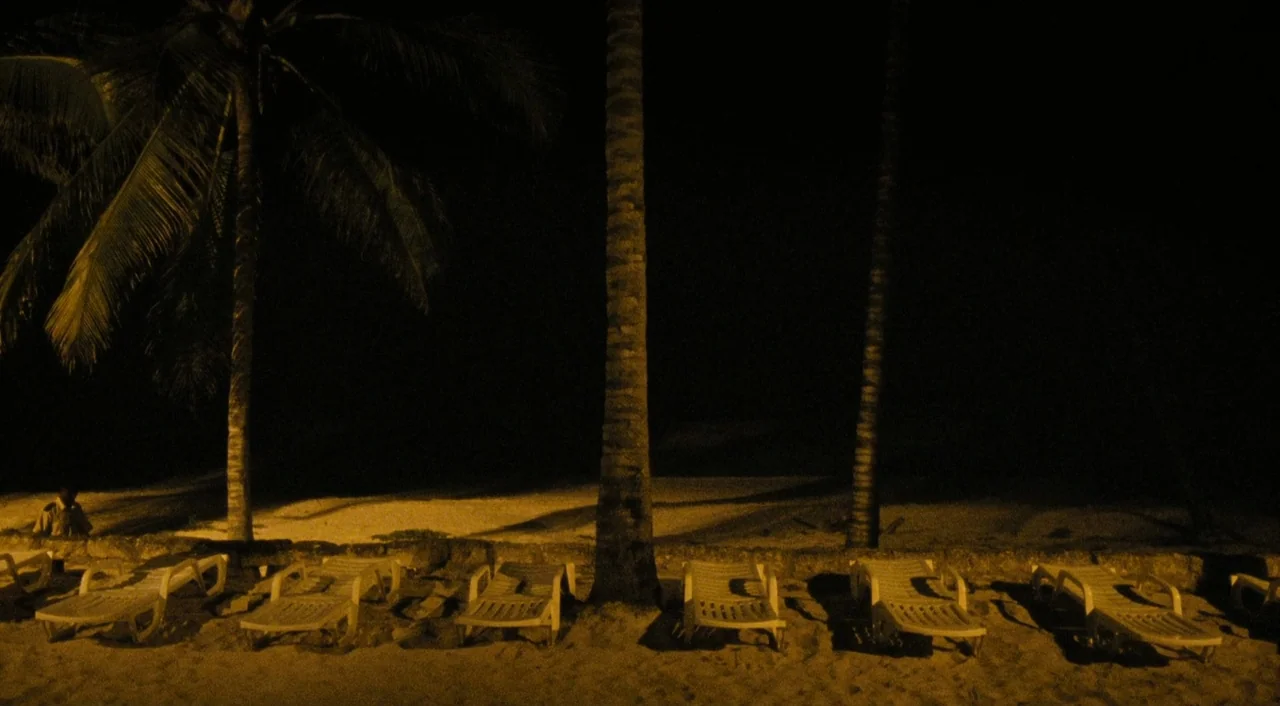 Paradise: Love (2012) movie still: wide — A dark beach area at night with several rows of white plastic lounge chairs. Three tall palm tree trunks…