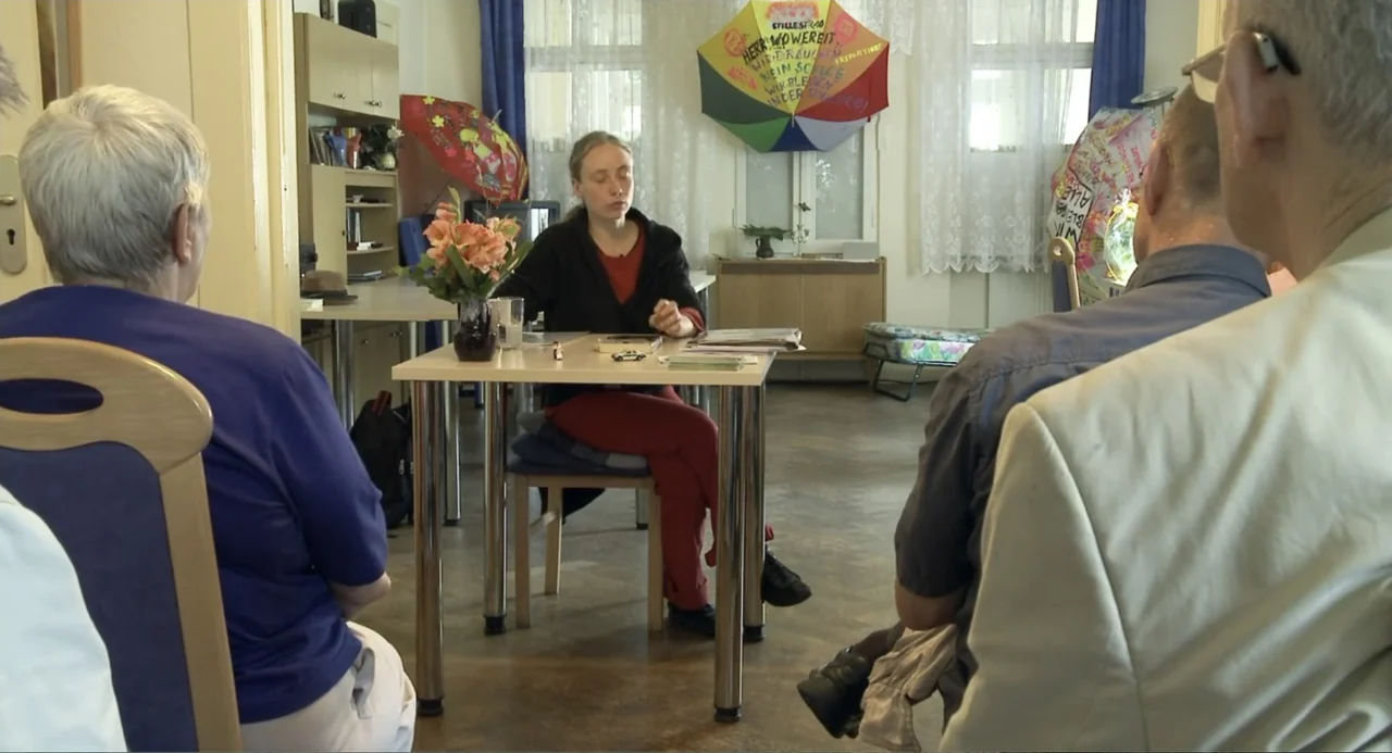 Az X- Kryptonit projekt (2020) movie still: medium — A functional communal room contains several tables and chairs with colorful decorations on…