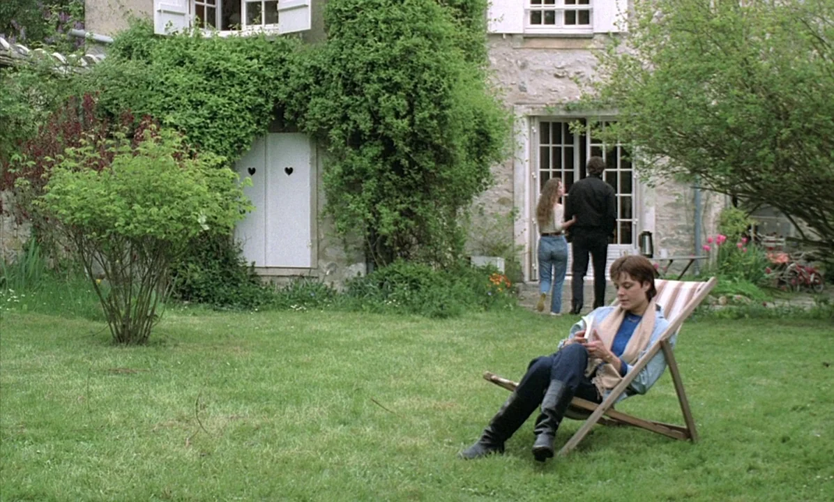 A Tale of Springtime (1990) movie still: wide — A stone country house with climbing ivy stands in a lush, grassy yard. A woman sits in a wooden deck…