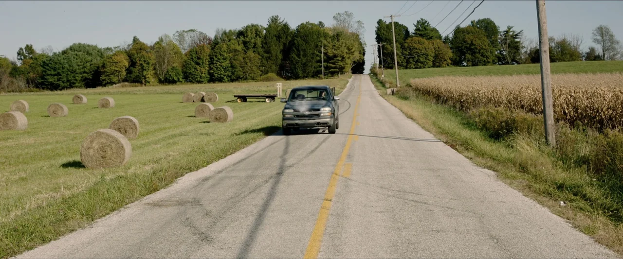 The Marksman (2021) movie still: wide — A rural asphalt road cuts through green fields with hay bales and corn crops. A grey pickup truck drives…