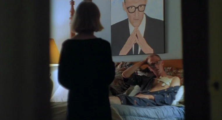 Schizopolis movie still: medium — A woman stands in a doorway looking into a dimly lit room with a painting. A man in a bathrobe rests on a bed with…