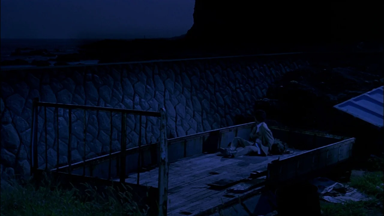 Battle Royale (2000) movie still: wide — A dark, nocturnal shoreline features a stone retaining wall behind an old metal truck. A young man sits…