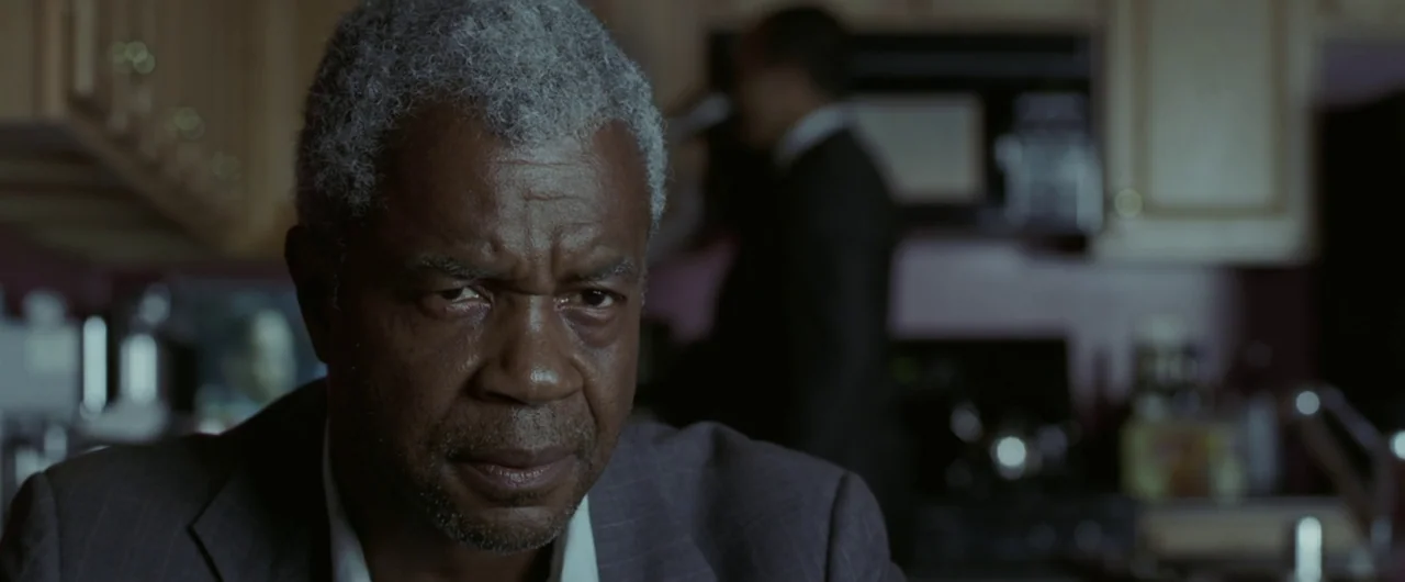 Syriana (2005) movie still: closeup — An older man stands in a dimly lit domestic kitchen with blurred appliances behind him. A second person in a…