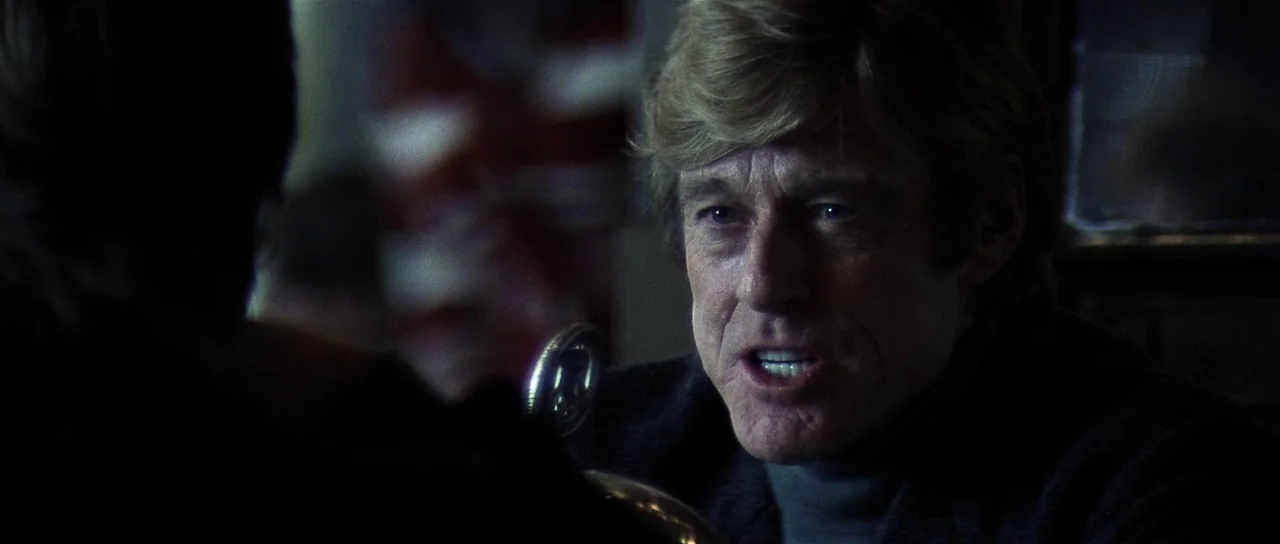 Spy Game (2001) movie still: closeup — A dark indoor space with a booth seating arrangement featuring dim practical lighting. An older man with…