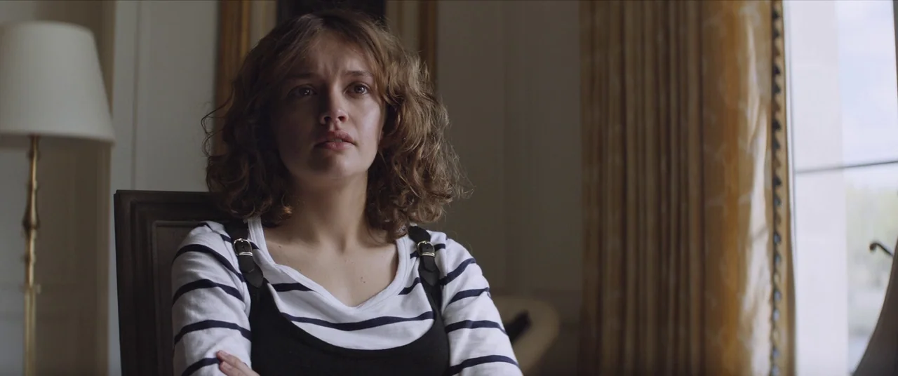 Thoroughbreds (2017) movie still: medium — A room with beige curtains and a tall floor lamp in the background. A woman in a striped shirt sits…