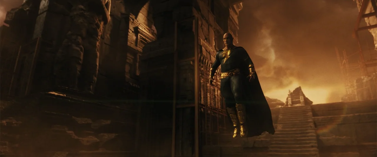 Black Adam (2022) movie still: fullbody — A crumbling ancient stone structure stands beneath a glowing sunset sky. A man in a dark costume with gold…