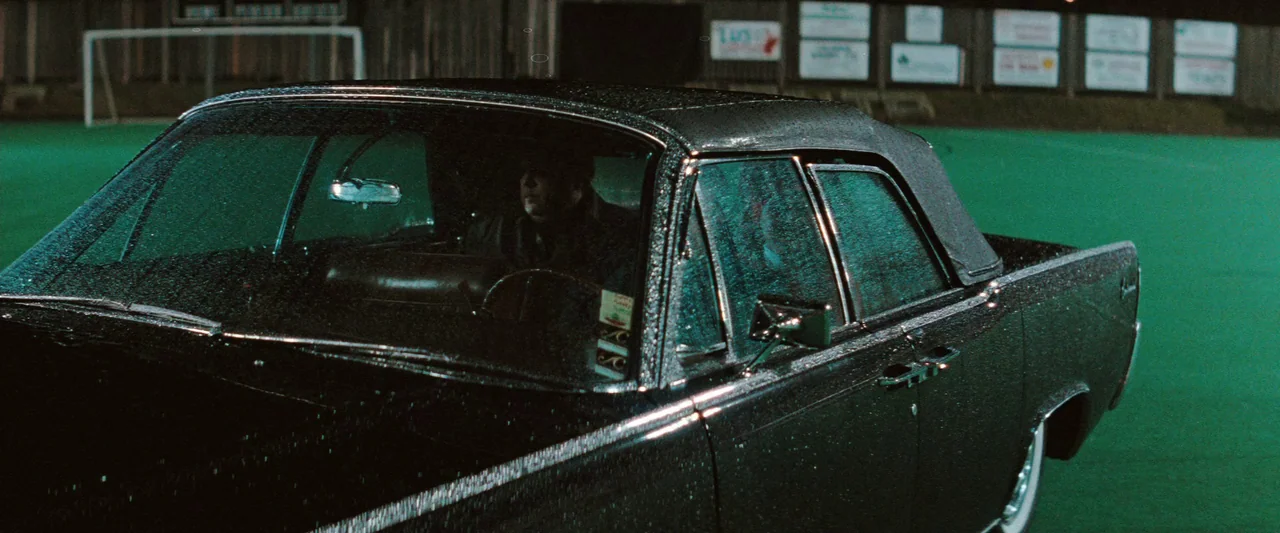 The Adjuster (1991) movie still: medium — A dark classic convertible sits on a wet, vibrant green artificial athletic field at night. An unseen…