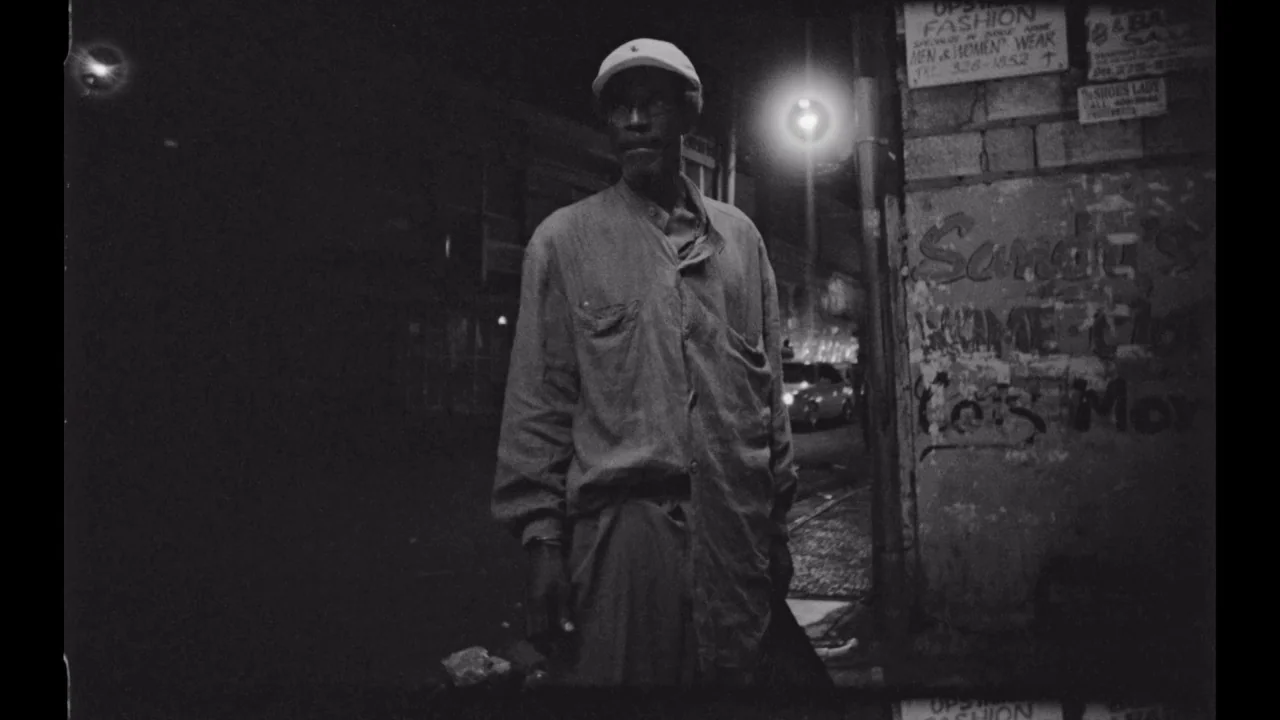 Black Mother (2018) movie still: fullbody — A dark city street corner featuring a brick wall with faded posters and graffiti. An elderly man stands…