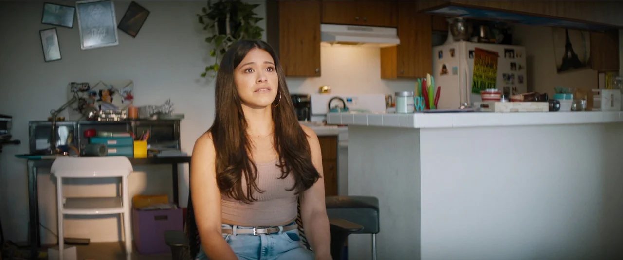 Kajillionaire (2020) movie still: medium — An apartment with a white kitchen counter and various personal items on the walls. A woman with long…