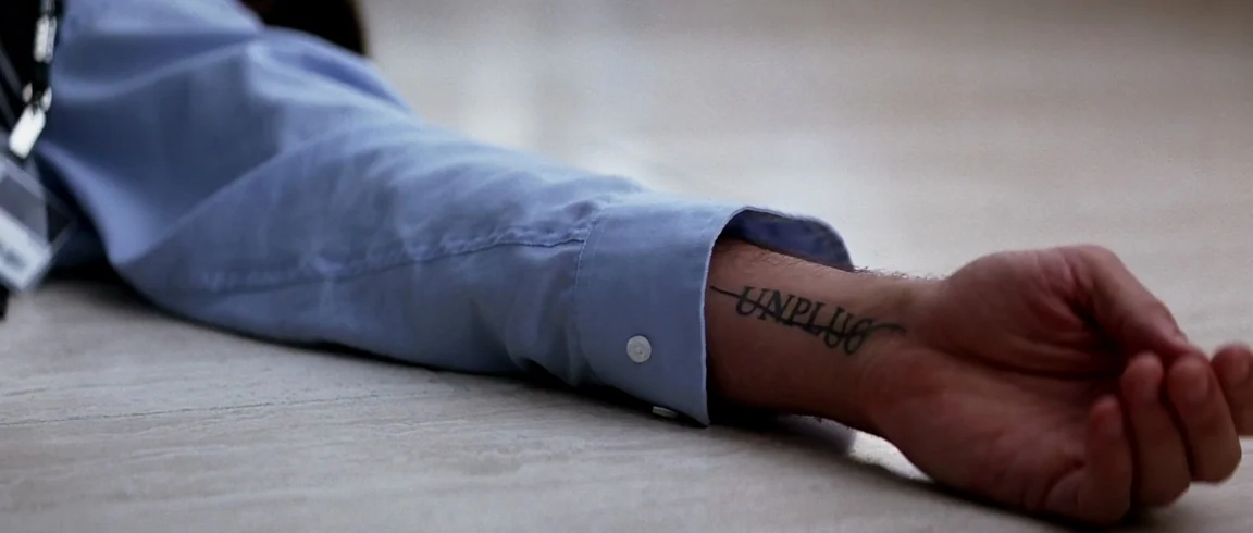 Transcendence (2014) movie still: closeup — A person lies with their arm stretched out on a light-colored tiled floor surface. The forearm displays…