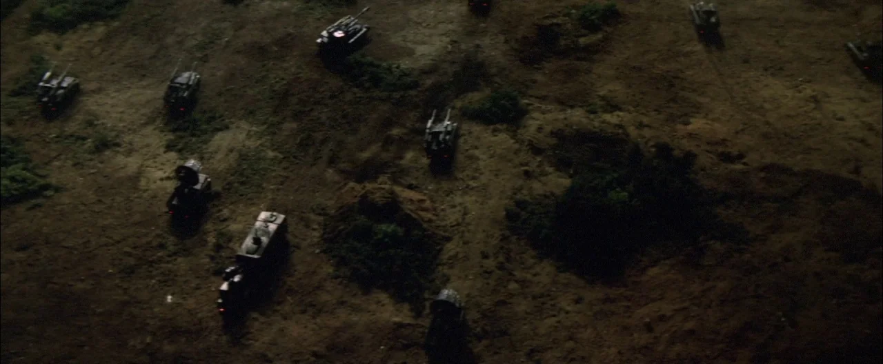 Destroy All Monsters (1968) movie still: establishing — A group of armored military vehicles traverses a dark, uneven patch of dirt. The uneven…