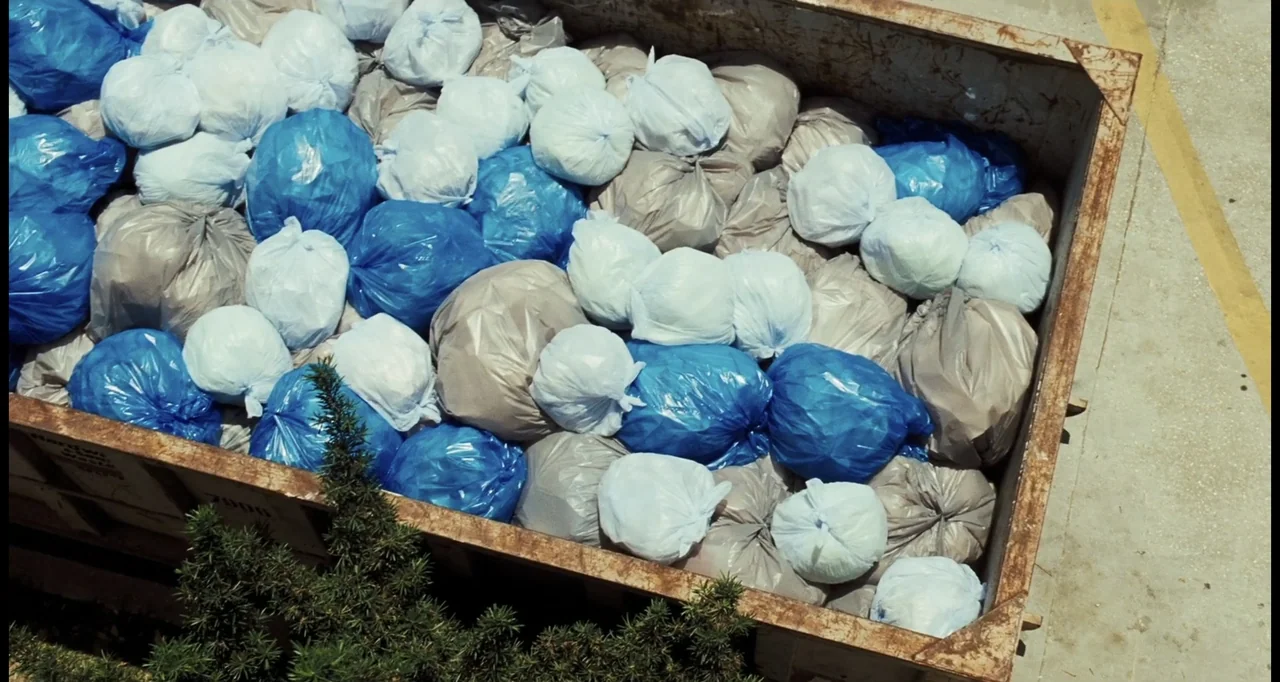 I Love You Phillip Morris (2010) movie still: wide — A metal industrial dumpster is filled with numerous blue, white, and grey trash bags. Green…