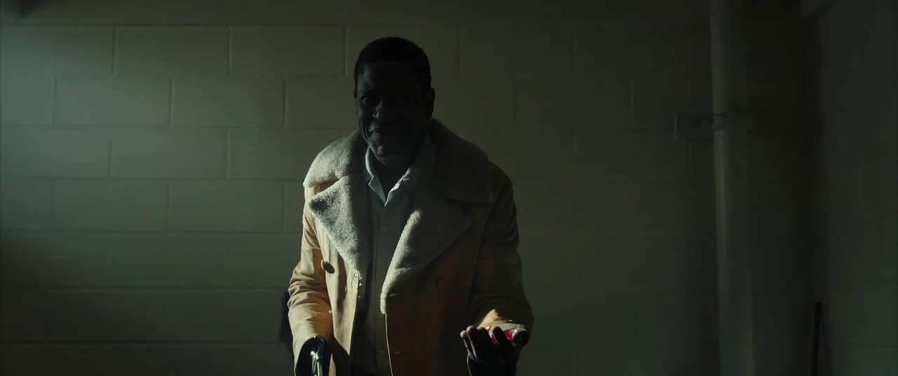 Candyman (2021) movie still: medium — A dimly lit basement room has walls constructed of unfinished concrete cinder blocks. An older man wearing a…