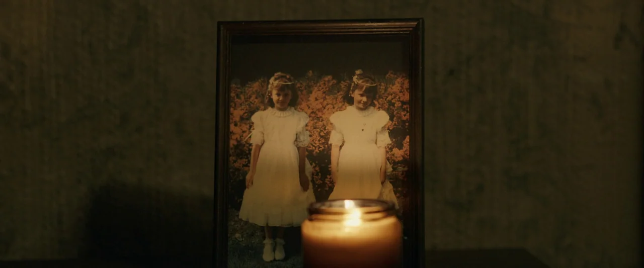 Oddity (2024) movie still: closeup — A dark, out-of-focus wall serves as the background for a framed portrait. The photograph depicts two identical…