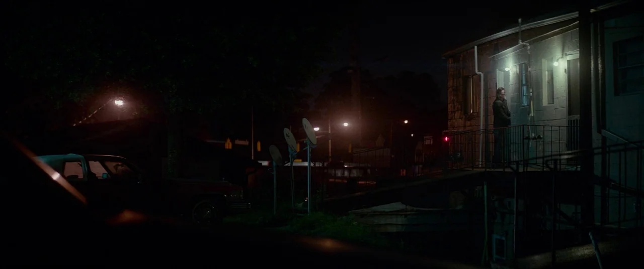 Triple 9 (2016) movie still: wide — A dark, low-rise concrete building stands at night with a metal walkway and ramp. A single man stands near the…
