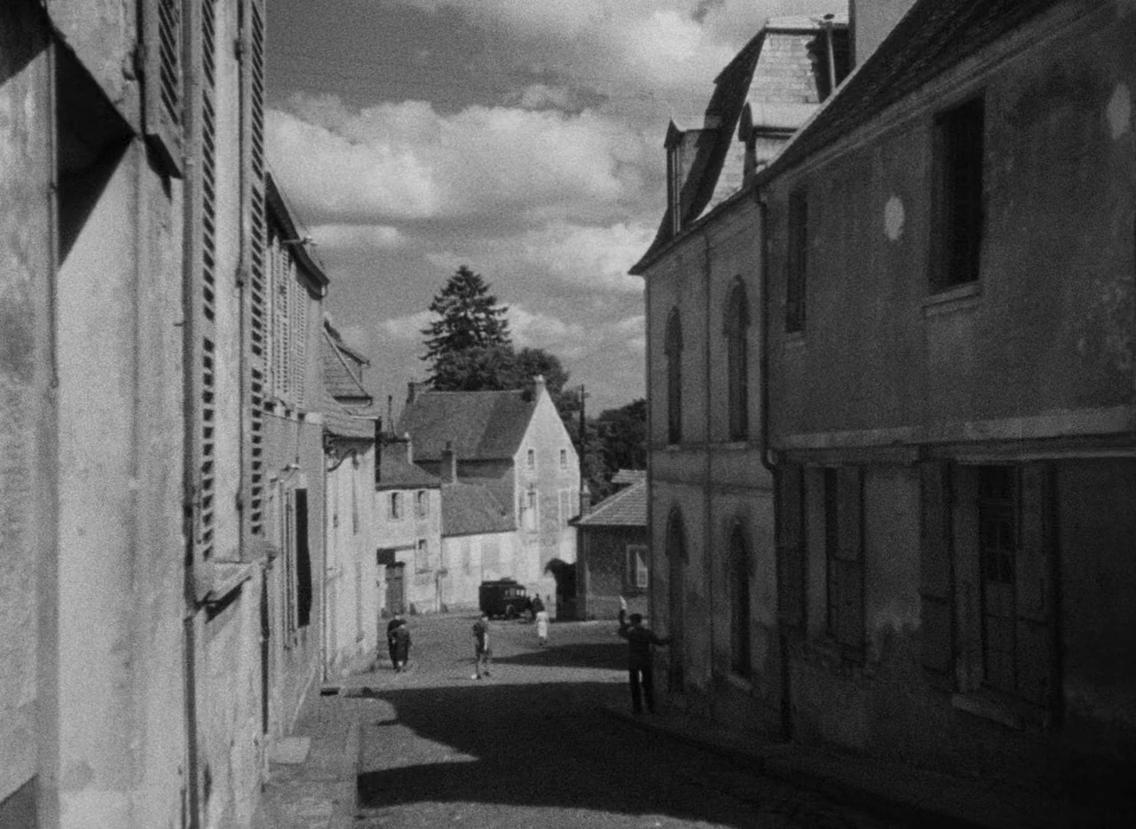Le Corbeau (1943) movie still: wide — A narrow European street is lined with weathered stone houses and closed wooden shutters. A few small figures…