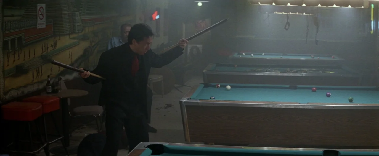 Rush Hour (1998) movie still: medium — A dimly lit billiard hall features multiple pool tables and a mural on the wall. A man in a suit brandishes…