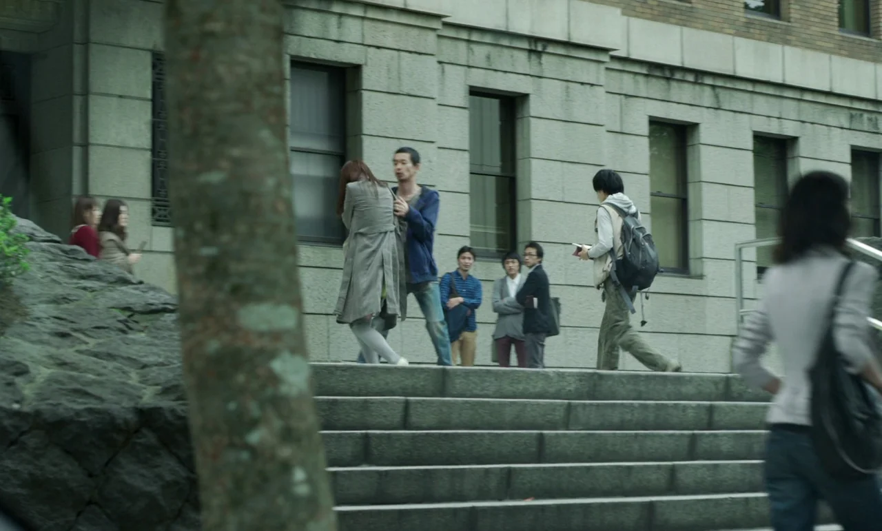 Like Someone In Love (2012) movie still: wide — A group of young adults congregates on wide stone steps in front of a building. A man holds the arm…