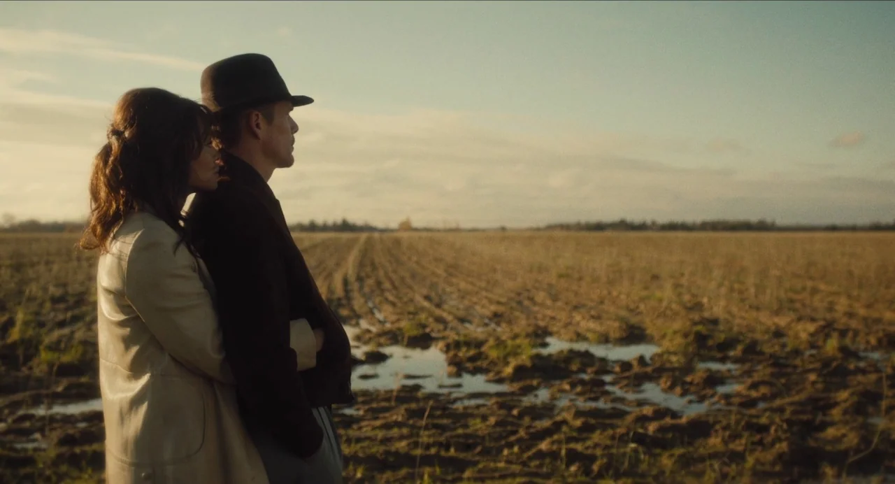 Born To Be Blue (2015) movie still: medium — A vast, muddy agricultural field stretches toward the horizon under a soft sky. A man in a hat and a…