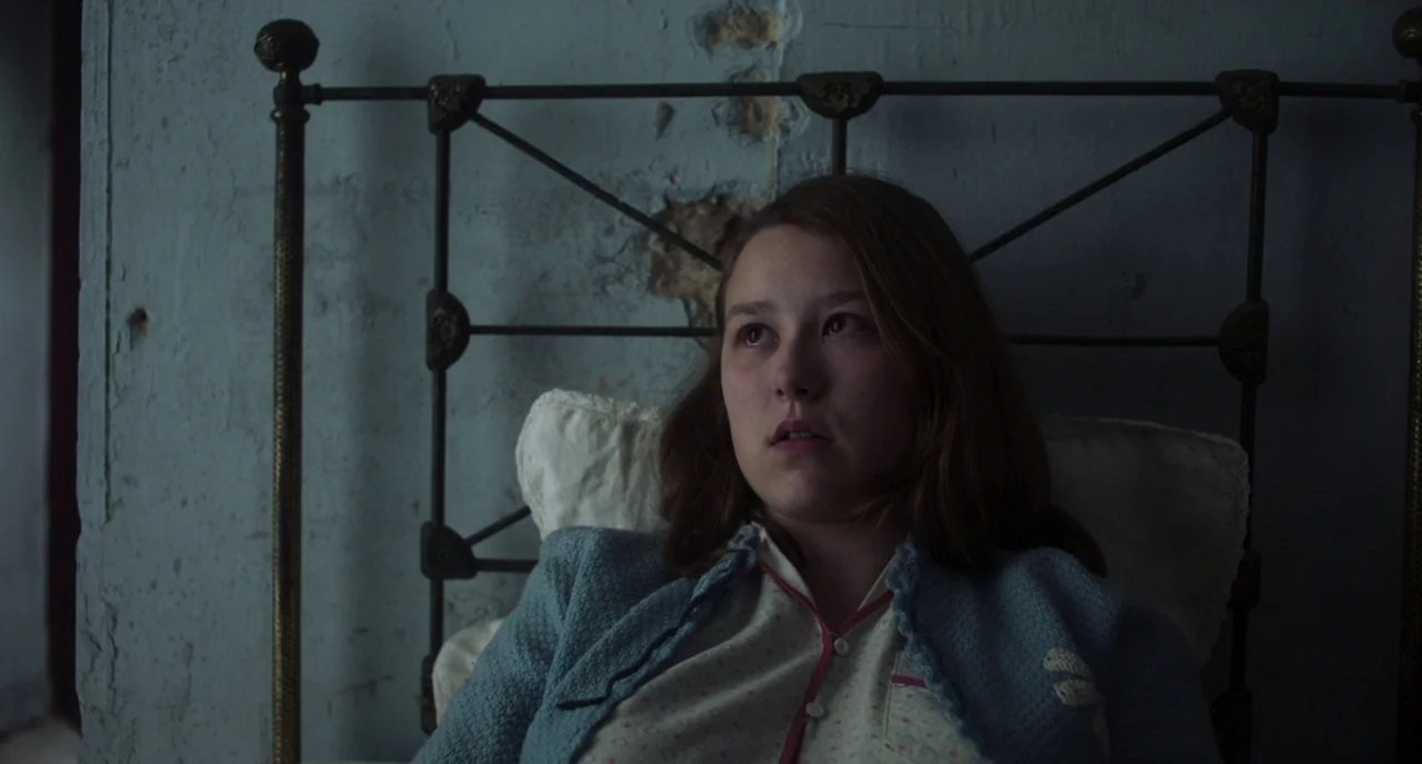 The Little Stranger (2018) movie still: medium — A dimly lit room contains an iron-framed bed against a weathered, peeling plaster wall. A teenage…
