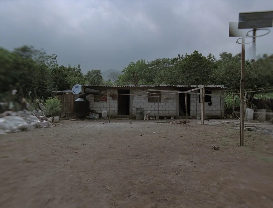 Post Tenebras Lux (2012) movie still: establishing — A low-slung concrete block building stands in a dusty rural clearing under grey clouds. Dense…