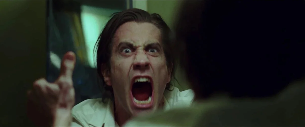 Nightcrawler (2014) movie still: closeup — A dimly lit room with muted green tones and a bright window in the background. A man screams directly…