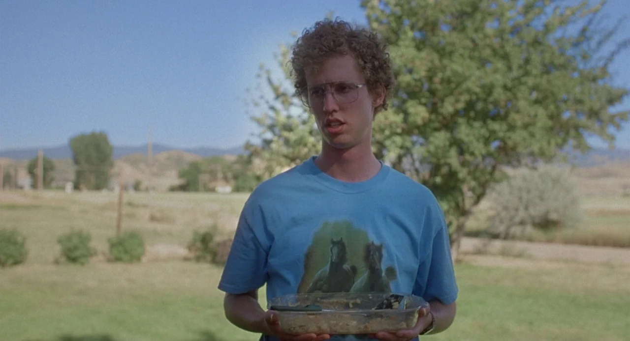 Napoleon Dynamite (2004) movie still: medium — A rural setting with a green lawn, sparse trees, and distant brown hills. A young man stands holding…