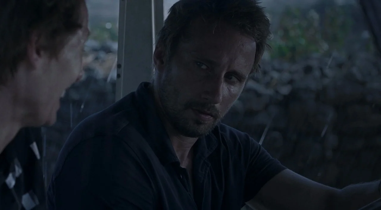 A Bigger Splash (2015) movie still: closeup — Two men sit inside a dimly lit vehicle during a rainstorm at night. The driver looks towards the…