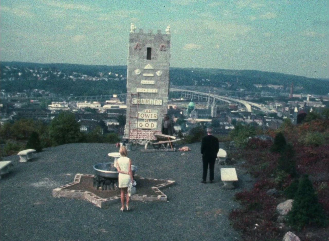Wanda (1970) movie still: wide — A gravel lookout features a central stone tower decorated with religious signage and text. A woman in a white dress…