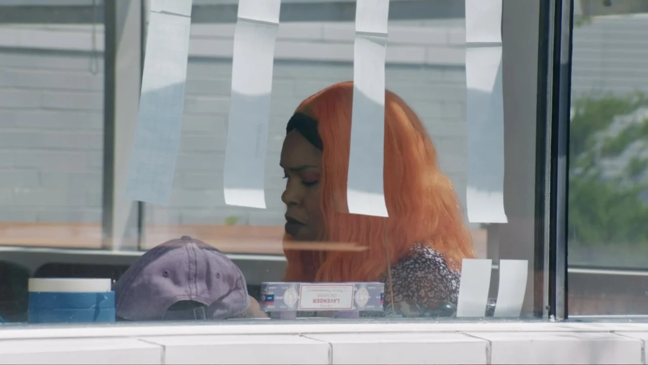 The African Desperate (2022) movie still: medium — A shop counter is viewed through a glass window with hanging paper strips. A woman with bright…