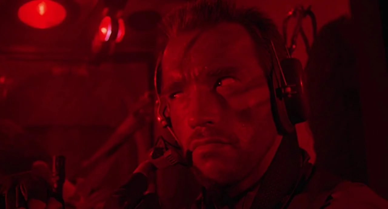 Predator (1987) movie still: closeup — An interior shot inside a transport helicopter bathed in deep red emergency light. A man in tactical gear…