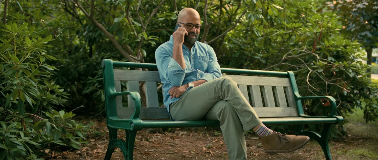 American Fiction (2023) movie still: medium — A man sits on a green metal bench surrounded by dense green shrubbery. The man is holding a phone to…