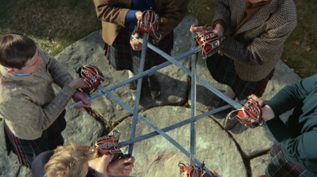 The Wicker Man movie still: wide — Five individuals stand in a circle on large, flat stone slabs outdoors. They hold swords in their hands, angling…