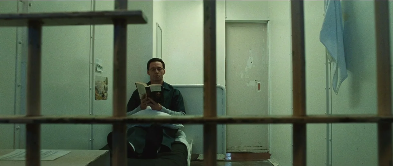 Bad Education (2019) movie still: medium — A spartan room features metal walls and a single bed with white linens. A man sits on the bed reading a…