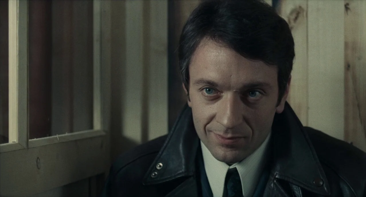 The Army of Shadows (1969) movie still: closeup — A man in a leather jacket stands inside a room with plain wooden paneling. The frame focuses on…