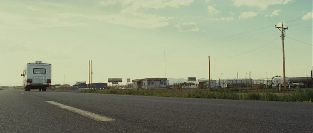 Paul (2011) movie still: wide — An open highway stretches into the distance beside a sparse, industrial roadside lot. A white recreational vehicle…