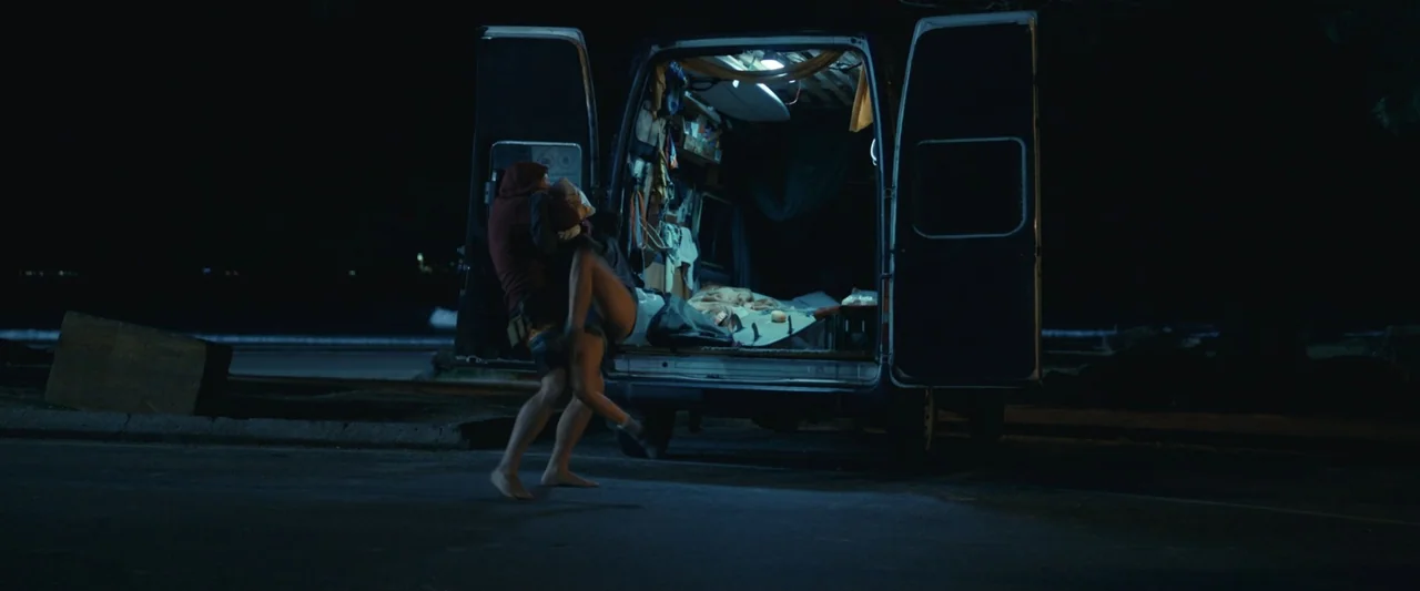 Dangerous Animals (2025) movie still: fullbody — A dark, paved area at night features a parked van with open rear doors. A man carries a woman…