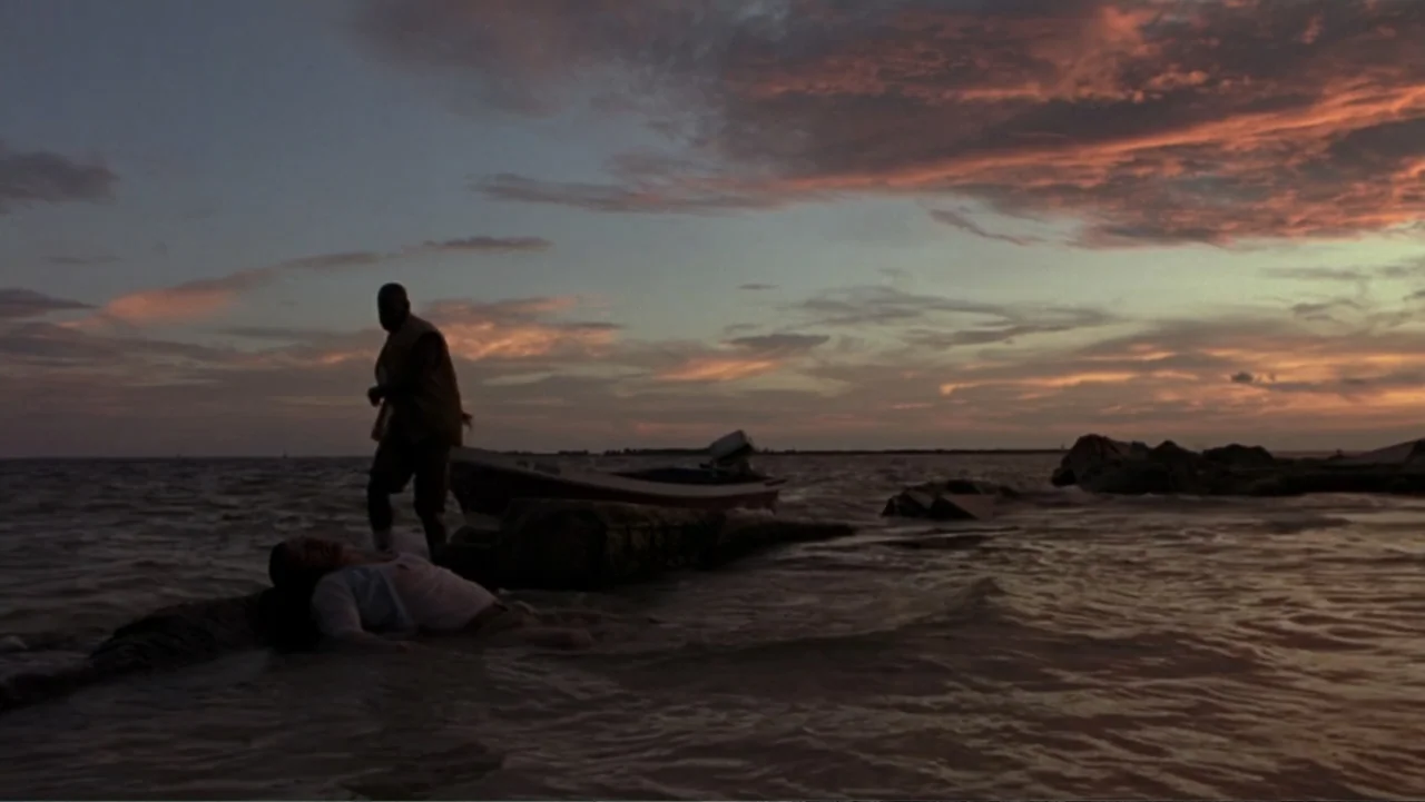 The Punisher (2004) movie still: wide — A rocky shoreline at golden hour with a small boat anchored in shallow waves. One man stands on the rocks…