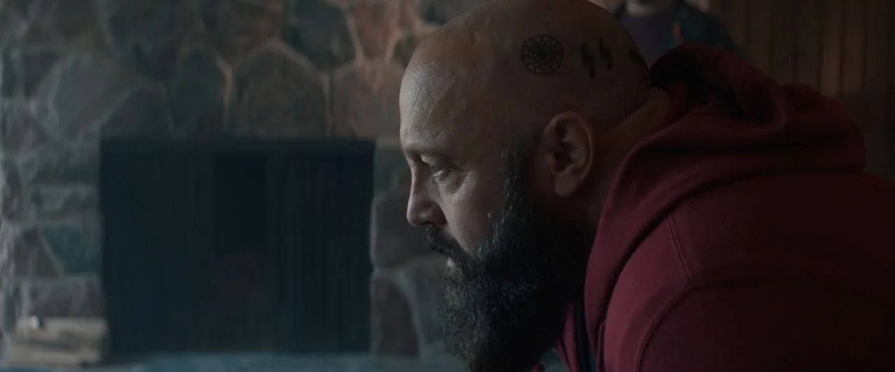 Becky (2020) movie still: closeup — A dark interior space features a rough-hewn stone fireplace backdrop. A bald man with facial tattoos and a dark…