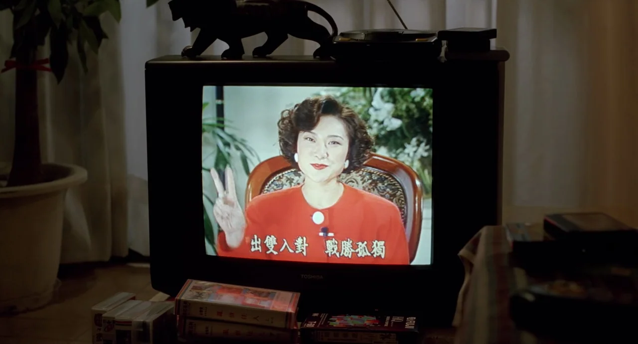 A Confucian Confusion (1994) movie still: medium — A television screen displays a woman speaking in a broadcast. Several VHS tapes are stacked in…