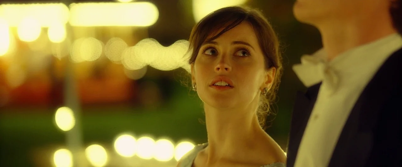 The Theory of Everything (2014) movie still: closeup — A woman stands in a nighttime fairground with bokeh lights glowing in the background. She…