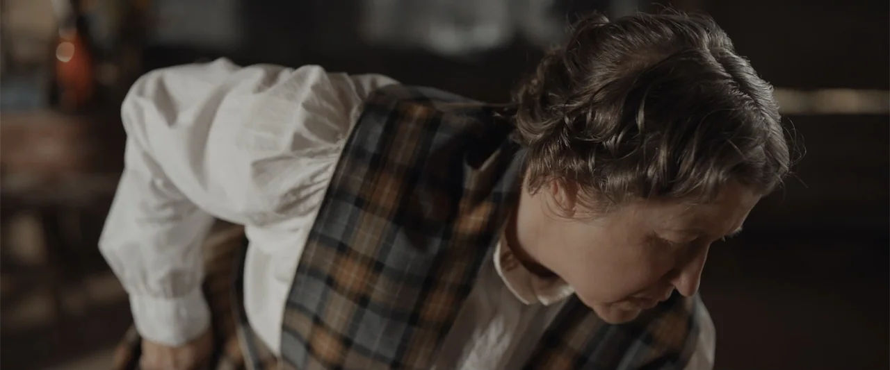Ghosts of the Ozarks (2022) movie still: medium — An interior space with a person dressed in 1900s style period clothing. The woman leans forward…