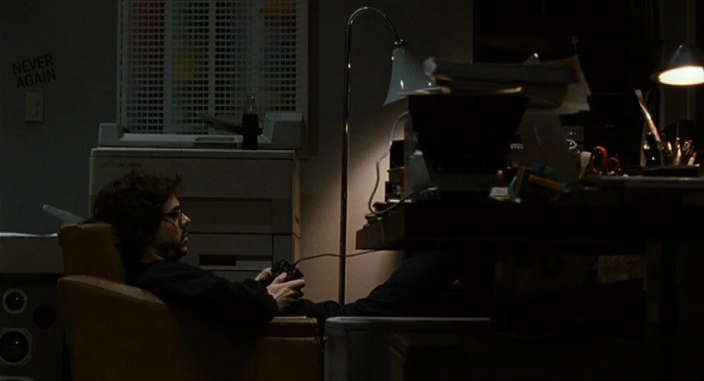 Sidewalls (2011) movie still: medium — A darkened home office features a cluttered desk and a large printer nearby. A man with glasses reclines in a…
