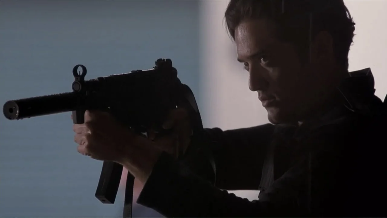 Crisis Point (2012) movie still: medium — A stark indoor setting with a plain light-colored wall visible behind the subject. A man in a dark jacket…