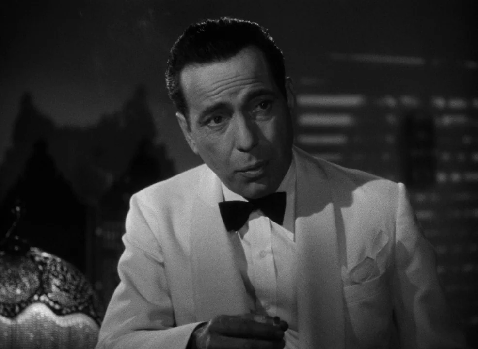 Casablanca (1942) movie still: medium — A man wearing a formal dinner jacket is inside a dimly lit room. The man holds a cigarette while looking at…