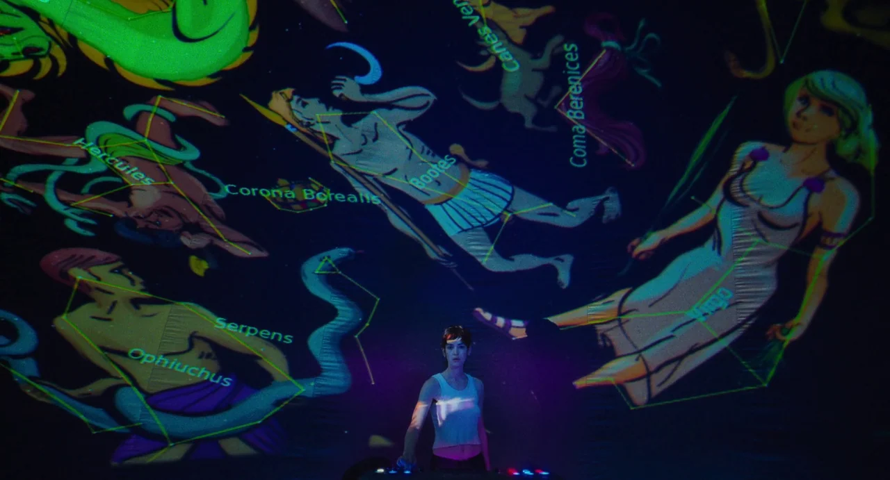 I Saw The TV Glow (2024) movie still: wide — A darkened room features a large projection of colorful constellation diagrams on the wall. A woman in…