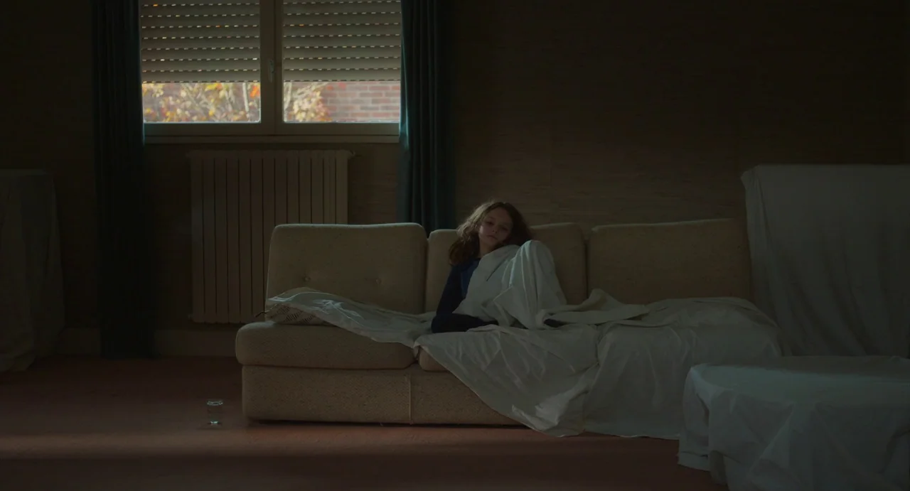 Petite Maman (2021) movie still: wide — A sparsely furnished room with a beige sofa covered by a white sheet. A girl sits on the sofa with a white…