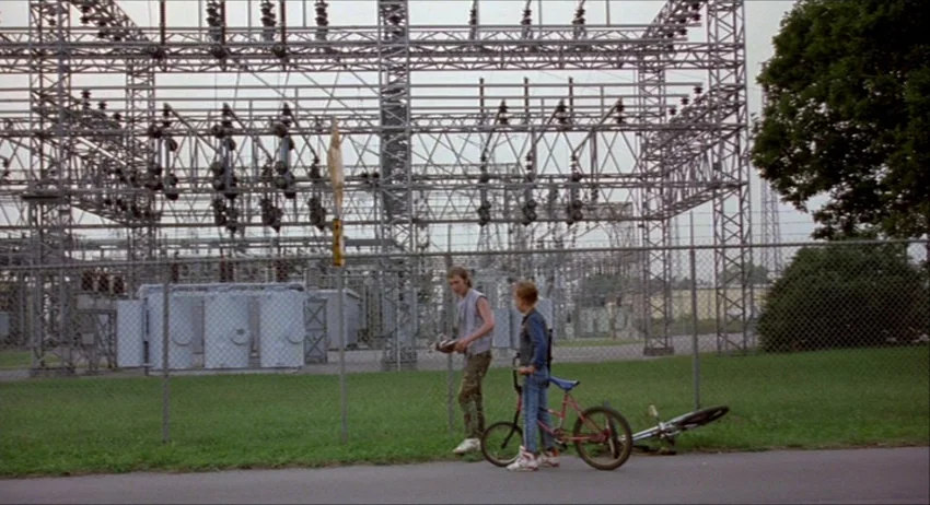 Gummo (1997) movie still: wide — An industrial electrical substation features a large metallic lattice structure behind a chain-link fence. Two boys…