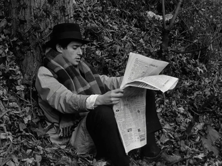 Band of Outsiders (Bande à part) (1964) movie still: medium — A man sits on the ground at the base of a tree in a woodland setting. He wears a…