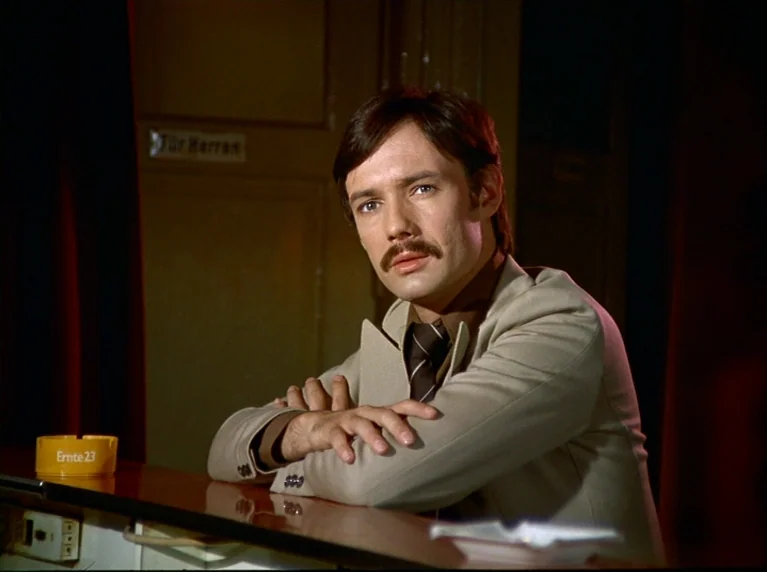 Fox and His Friends (1975) movie still: medium — A man sits at a dark bar counter against a blurred wooden door background. The man with a mustache…